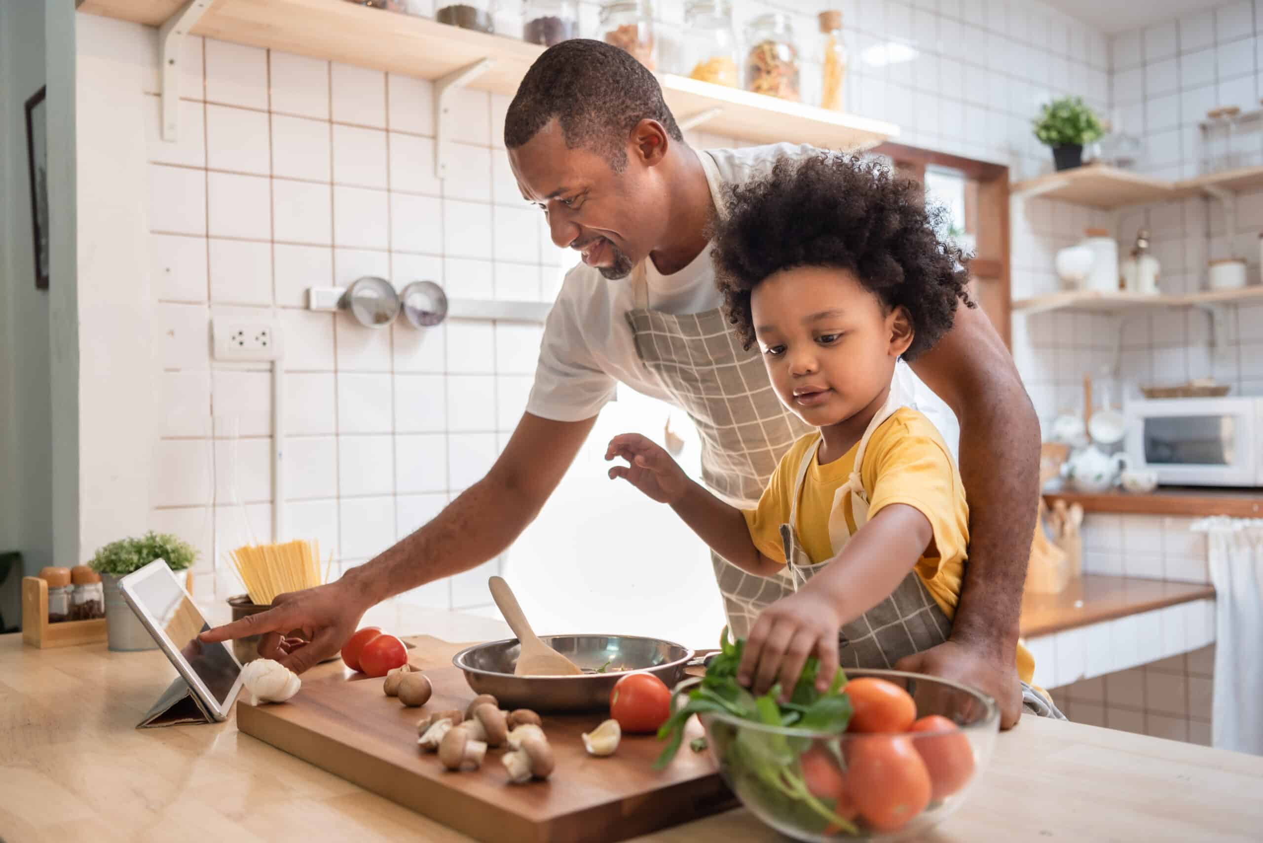 African American Little boy preparing food while his father looking on the digital recipe and using touch screen tablet in the kitchen at home